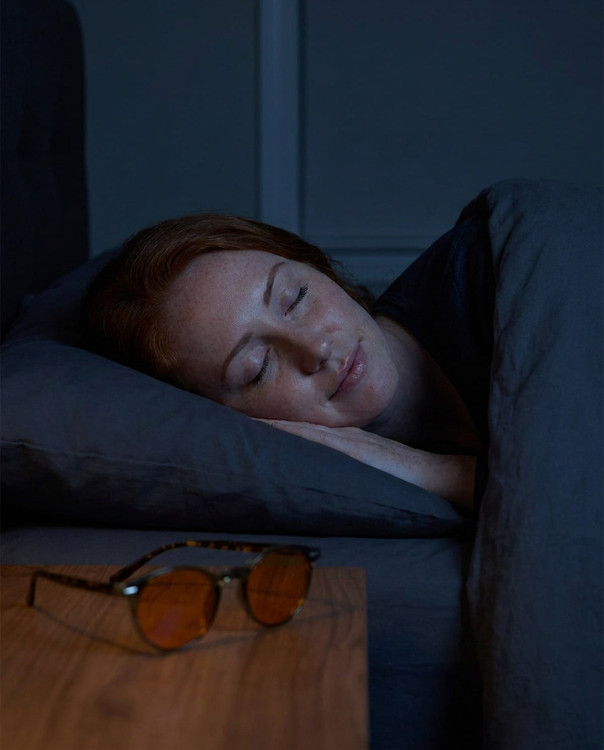 Illustration: woman asleep in a dark bedroom, Seluna amber glasses folded on the nightstand