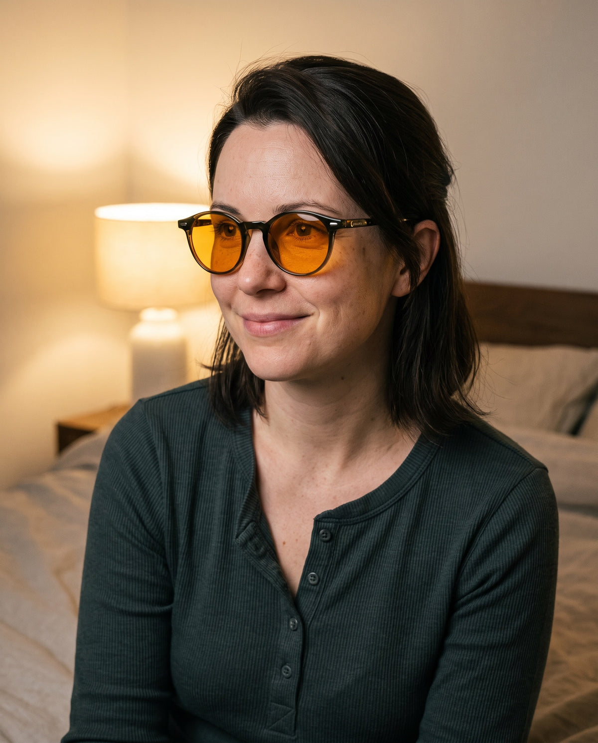 Woman with dark hair wearing Seluna amber glasses in a green henley on her bed in the evening